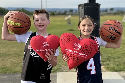 Jameson Shaw and Odin Schneider smiling while holding basketballs and red Seattle Children's Hospital heart pillows.