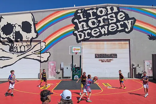 Kids playing a 3-on-3 basketball game on an outdoor court set up in front of the Iron Horse Brewery building mural.